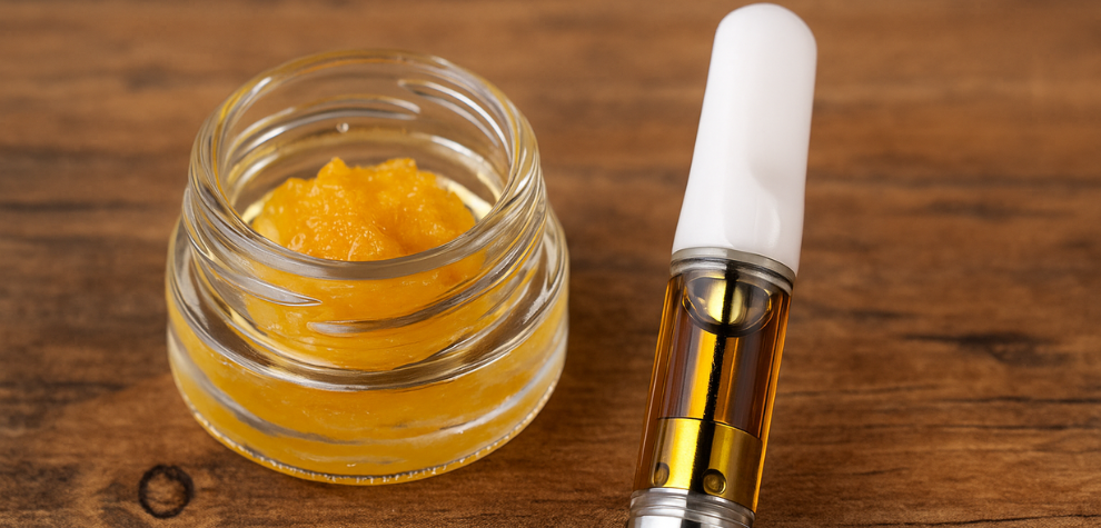 A glass jar of golden live resin next to a filled vape cartridge with a white mouthpiece on a wooden surface